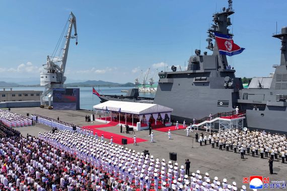 North Korean leader Kim Jong Un stands at a podium addressing a naval commissioning ceremony, with a large grey warship behind him.