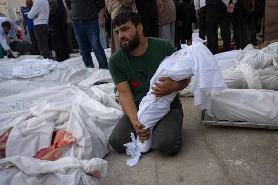 A man holds a small body bag next to other bodies in body bags on the ground.
