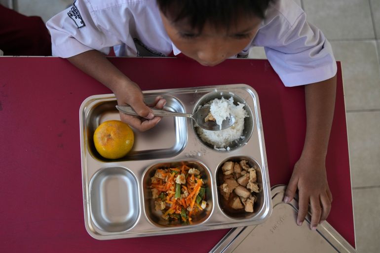 A young student in school uniform eats from a meal tray at a classroom desk, with other children eating around him